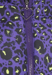 Fialová nadýchaná bunda s černým a neonově žlutým leopardím potiskem. Má centrální zip s přívěskem ve tvaru srdce a prošívanou texturu.