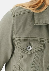 Veste en denim olive avec un col structuré, une poche poitrine avant et des détails en boutons-pression argentés. Tissu texturé et couture visible.