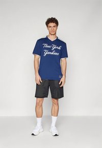 Tamsiai mėlynas V formos megztinis su užrašu „New York Yankees“ baltu šriftu. Derinamas su juodomis tinklelio šortais ir baltais sportbačiais. Lygios audinio tekstūros.