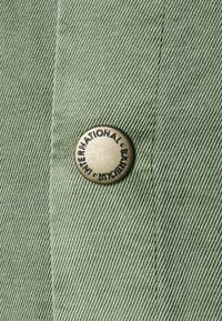 Grüner Stoff mit diagonaler Textur, versehen mit einem runden Metallknopf, auf dem "INTERNATIONAL BARBARA" eingraviert ist.