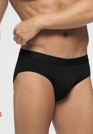 Primo piano del torso maschile che indossa slip neri, mostrando l'addome inferiore, i fianchi e la parte superiore delle cosce su sfondo uniforme.