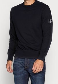 Svart sweatshirt med långa ärmar, rund halsringning och ribbade muddar. Har en logotypmärke på vänster ärm. Bärs med blå jeans.