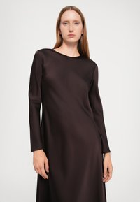 Langermet, mørkebrun kjole laget av glatt stoff. Har en rund halslinje og en løs passform, med elegante linjer og minimal pynt.