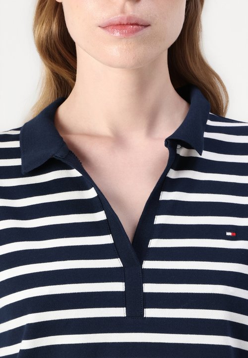Polo a righe blu navy e bianche con colletto e scollatura a V, caratterizzato da un piccolo logo sul lato sinistro del petto. Il tessuto sembra strutturato.