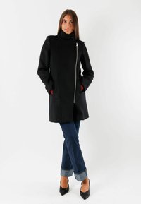 Cappotto di lana nero con collo alto, zip frontale e tasche laterali. Interno rosso a contrasto. Indossato con jeans blu e décolleté nere a punta.