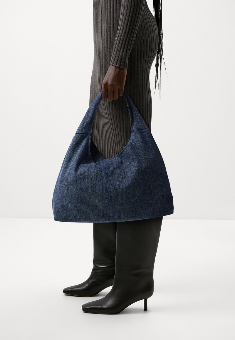 Personne en robe grise côtelée et bottes noires à talons tenant un grand sac à main en tissu bleu texturé par sa poignée sur le côté.