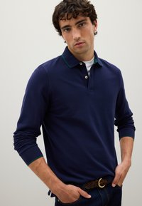 Mann mit lockigem, dunklem Haar, der ein langärmliges Poloshirt in Marineblau mit grünem Besatz und einen braunen geflochtenen Gürtel trägt, vor einem einfarbigen Hintergrund stehend.