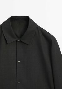 Manteau noir boutonné avec un large col et des boutons-pression mats, vu de dessus sur fond blanc.