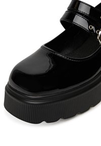 Zapato Mary Jane de charol negro con puntera redondeada, doble correa, herrajes plateados y suela gruesa texturizada.