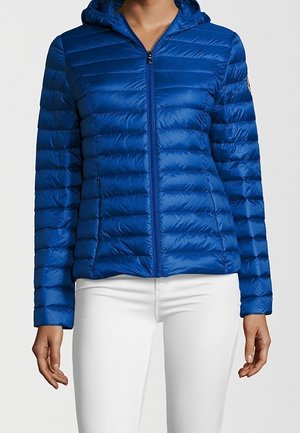 Mujer con una chaqueta ajustada acolchada y brillante de color azul con capucha y cremallera, y pantalones blancos, de pie contra un fondo neutro.