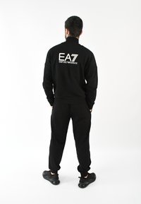Chándal deportivo negro con cuello alto, que presenta el logo "EA7 Emporio Armani" en la parte posterior. El material parece suave, con un ajuste holgado.