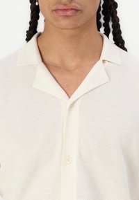 Knit cream polo shirt with a textured finish, flat collar, and a single button closure at the neck. Short-sleeved design details visible.