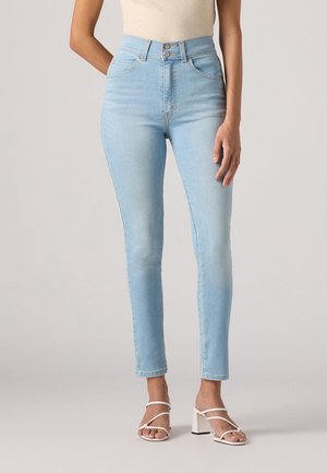 Frau trägt hellblaue High-Waist Skinny Jeans in Kombination mit weißen Riemchen-Blockabsatz-Sandalen, steht vor einfarbigem Hintergrund.