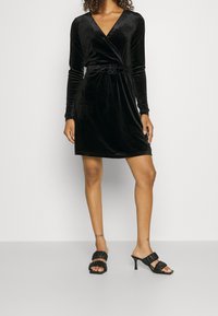 Svart flöjl omfångsdräkt med långa ärmar och bältad midja; jämn textur, över-knä längd; i kombination med svarta klacksandaler.