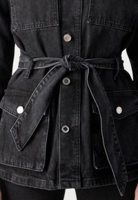 Črna jeans jakna z zavezanim pasom, srebrnimi pritiski in dvema sprednjima žepoma. Ima strukturirano zasnovo in vidne šive.