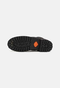 Suela de bota negra con surcos profundos, que presenta un acento de logo naranja. Material de goma texturizada con clavos metálicos para mayor agarre.
