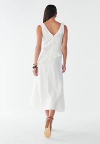 Weißes Midikleid mit Rüschendetails entlang des Rückens und der Schulter. Hat einen V-förmigen Ausschnitt und strukturierte Stoff. Enthält Riemchenschuhe.
