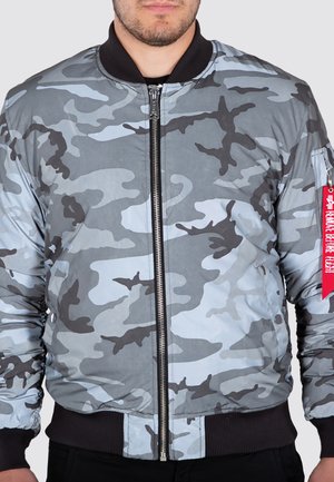 Mann trägt eine Bomberjacke in Grau und Schwarz mit Tarnmuster, rotem Etikett am linken Ärmel sowie schwarzem Kragen und Bündchen.