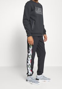 Sweat-shirt noir avec poche zippée et logo graphique. Pantalon noir avec motif abstrait coloré sur le côté. Chaussures de sport grises.