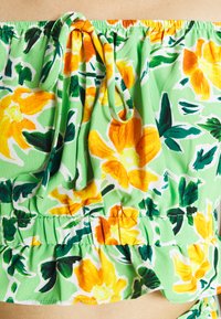 Top floreale off-shoulder verde con fiori gialli e arancioni. Presenta un volant elastico, un dettaglio con lacci e un tessuto leggero e testurizzato.