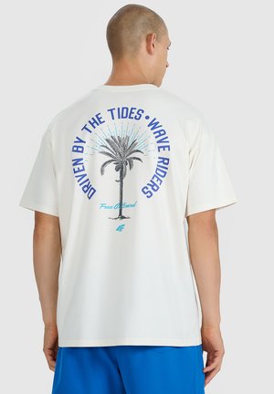 Mężczyzna w białej koszulce z grafiką palmy i napisem „Driven by the tides, wave riders, Free On Board“ na plecach, w połączeniu z niebieskimi szortami.