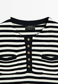 Randig svart-och-vit bomullstopp med rund halsringning, fem guldfärgade knappar och två fickdetaljer fram.