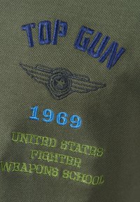 Tkanina w kolorze oliwkowym z haftowanym napisem: "TOP GUN" w niebieskim, "1969" w jasnoniebieskim oraz "UNITED STATES FIGHTER WEAPONS SCHOOL" w zielonym.