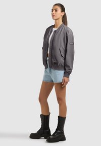 Femme portant une veste bomber grise, un crop top blanc, un short en denim clair et des bottes de combat noires, debout devant un fond clair uni.