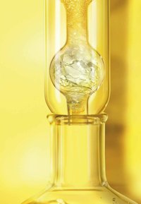 Bouteille en verre transparent avec un sommet rond et un col étroit, contenant un liquide doré avec des bulles et un élément sphérique à l'intérieur. Fond jaune.