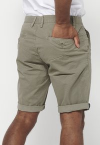 Man in beige opgerolde shorts met hand in achterzak, tegen een effen lichte achtergrond.