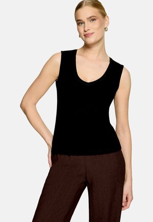 Femme aux cheveux blonds portant un haut noir sans manches à col en V et un pantalon texturé marron foncé, posant avec une main sur la hanche devant un fond uni.