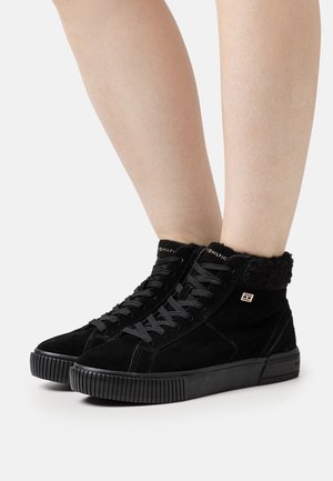 Czarne wysokie trampki z futrzanym podszyciem i sznurowadłami, noszone na gołych nogach, z małym złotym logo po boku i prążkowanymi gumowymi podeszwami.