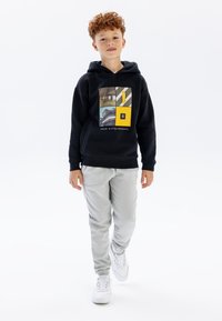 Sort hættejakke med grafisk tryk, grå sweatpants og hvide sneakers. Hættejakken har et farverigt design med firkanter og en gul accent.