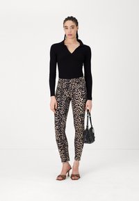 Maglietta nera a maniche lunghe a coste, pantaloni aderenti a stampa leopardata, tacchi con cinturini trasparenti, in mano una piccola borsa nera. Sfondo bianco semplice.