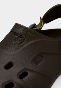 Κοντινό πλάνο σκούρου καφέ παπουτσιού Crocs που δείχνει υφή στην επιφάνεια, οπές εξαερισμού και ρυθμιζόμενο λουράκι στη φτέρνα.