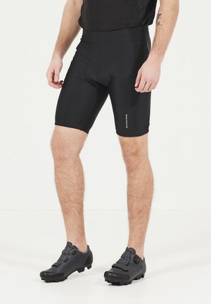 Schwarze Kompressionsshorts mit glatter Textur, ausgestattet mit einem gepolsterten Einsatz und einem engen Schnitt. Dazu schwarze Radschuhe mit strapazierfähigen Sohlen.