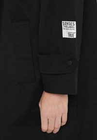 Nahaufnahme einer Hand neben einem schwarzen Ärmelsaum mit einem Druckknopfabschluss und einem weißen Patch, der "SENSES THE LABEL TRUE" beschriftet ist.