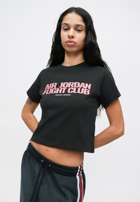 Νεαρή γυναίκα με μακριά μαύρα μαλλιά, φορώντας ένα μαύρο κοντό μπλουζάκι με ροζ κείμενο "Air Jordan Flight Club" και σκούρες φόρμες με πλευρικές ρίγες.