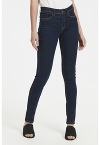 Jeans ajustados de mezclilla azul oscuro, con una cintura de altura media, diseño de cinco bolsillos y detalles de costura sutiles. Llevados con zapatos negros de abertura en los dedos.