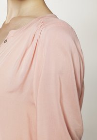 Světle růžová blůza s jemnou texturou, kulatým výstřihem a jemnými sklady na ramenou. Obsahuje malý knoflíkový detail.