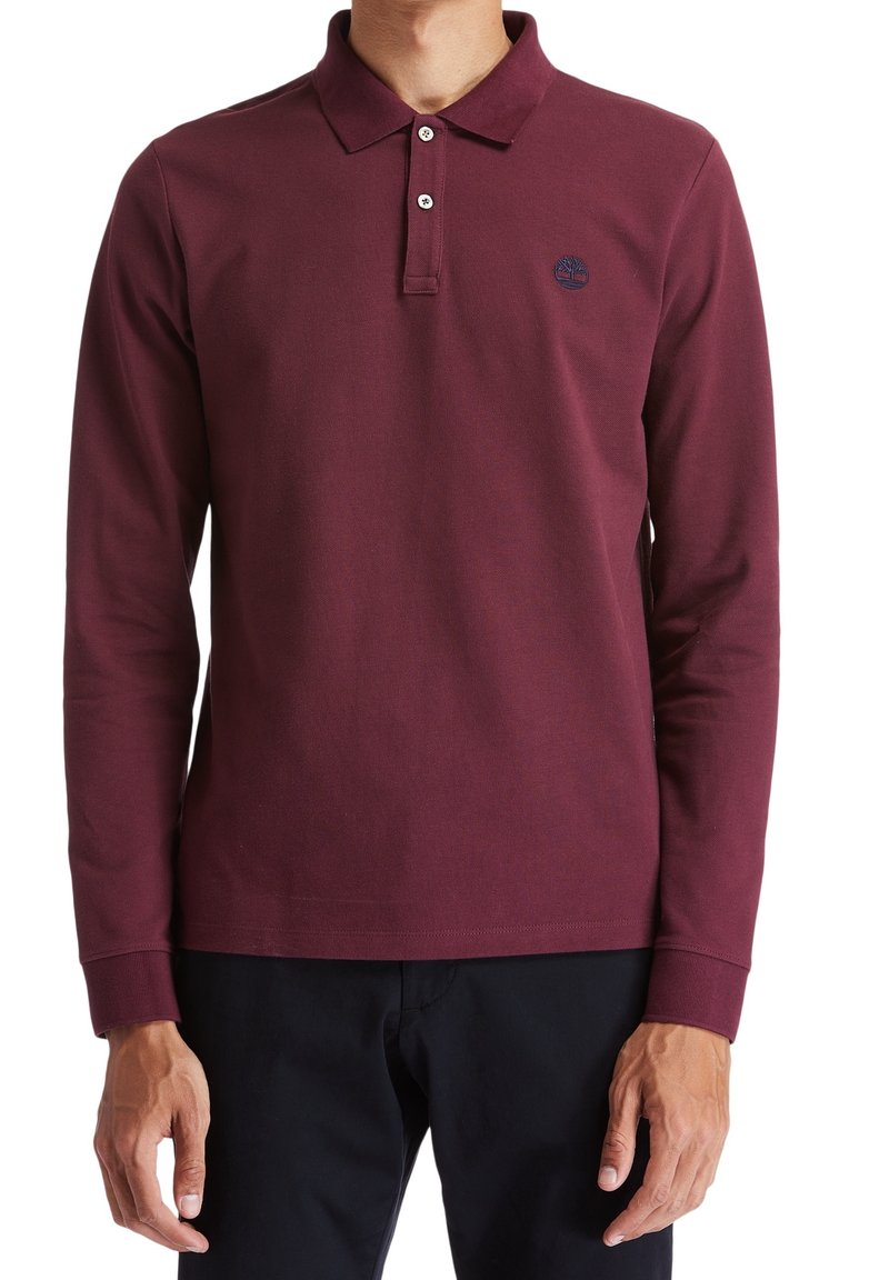 Homme portant un polo bordeaux à manches longues avec boutons et un pantalon noir, debout devant un fond uni.