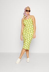 Vestido amarelo até ao joelho com padrão floral, alças finas e design ajustado. Combinado com sapatos brancos tipo slip-on e um lenço rosa.