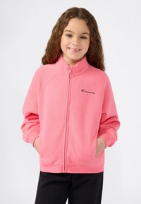 Ragazza sorridente con lunghi capelli ricci che indossa una felpa rosa con zip e ha le mani nelle tasche, su uno sfondo semplice.