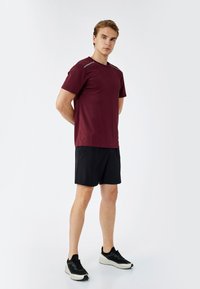 Camicetta corta color marrone con accenti grigi, abbinata a pantaloni corti neri. Il modello indossa scarpe da ginnastica nere, su uno sfondo bianco semplice.