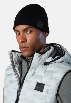 Cappello a maglia nero e giacca grigia con zip, indossati sotto un gilet imbottito bianco con motivo strutturato e patch logo sul petto sinistro.
