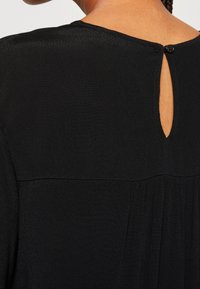 Blusa negra con abertura en la espalda y cierre de botón, textura suave y pliegues sutiles en la parte posterior para un interés visual añadido.