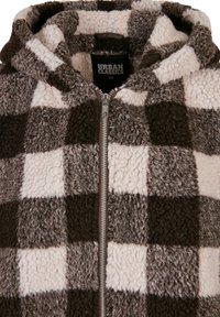 Vilnos striukė su gobtuvu ir užtrauktuku, pasižyminti rudo ir kreminio spalvų langelių raštu bei minkšta, tekstūruota medžiaga. Viduje matoma prekės ženklo etiketė.