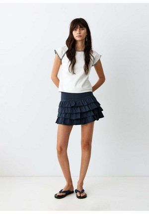 Frau mit langen dunklen Haaren, die ein weißes ärmelloses Oberteil mit schwarzem Saum, einen marineblauen gestuften Rock und schwarze Flip-Flops trägt, vor weißem Hintergrund stehend.