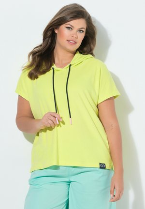 Kobieta z długimi brązowymi włosami, ubrana w jasnżółtą bluzę z krótkim rękawem i miętowe spodnie, stojąca na białym tle.