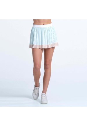 Frau trägt einen plissierten hellblauen Minirock mit beigem Saum und weißen Turnschuhen, steht mit einem Fuß leicht nach vorne.